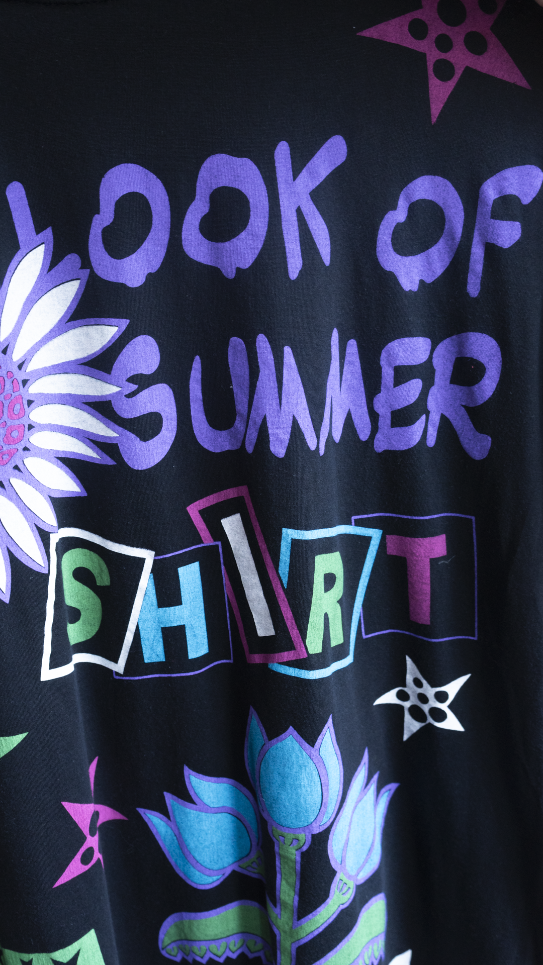 detail of 80s oversized vintage t-shirt dress in black with colorful graphics and text 'LOOK OF SUMMER SHIRT' in purple, blue, and white retro-style lettering, decorated with purple and blue flowers and star motifs.