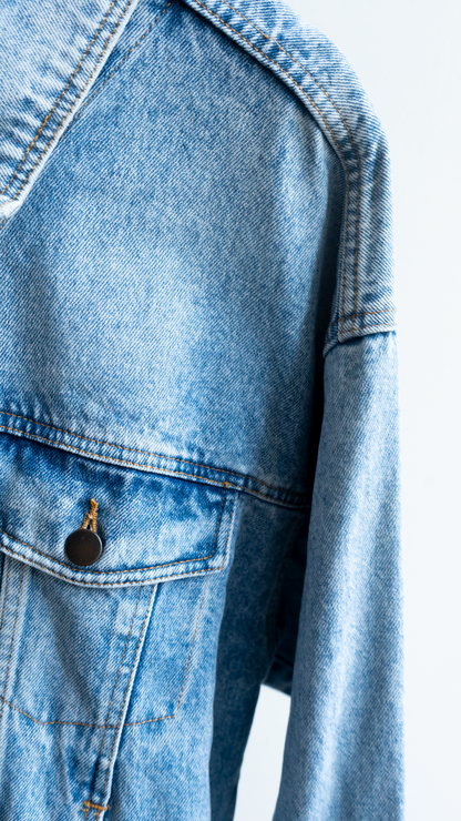 Close-up of a vintage 90s light blue denim jacket showing the chest pocket with a dark metal snap button and the textured denim fabric