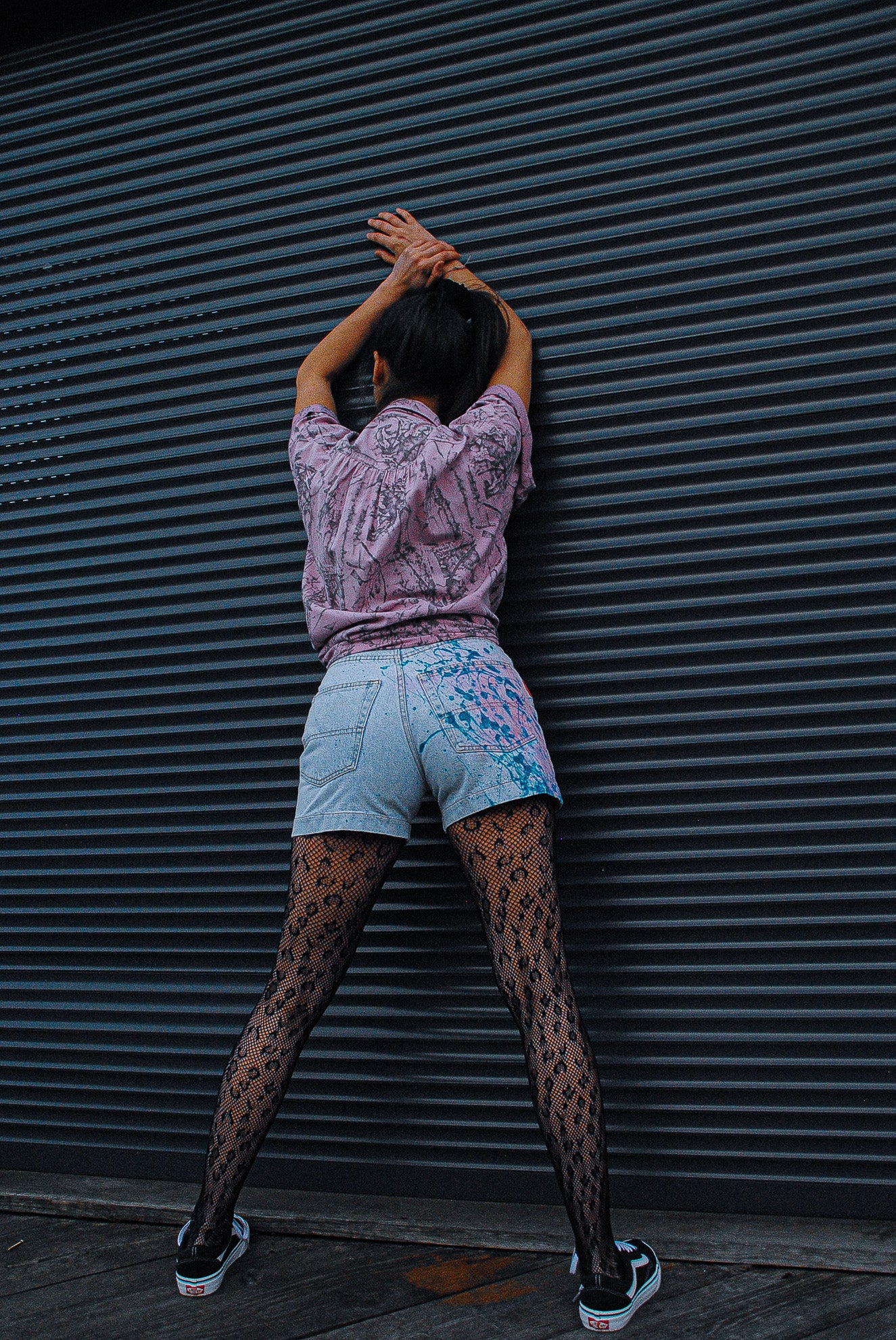 model-wearing-leopard-stockings-and-light-jeans-short-with-paint-splatters-in-blue-and-pink-on-the-back-from-the-fresh-vintage-art-wear-collection-leaning-towards-garage-door