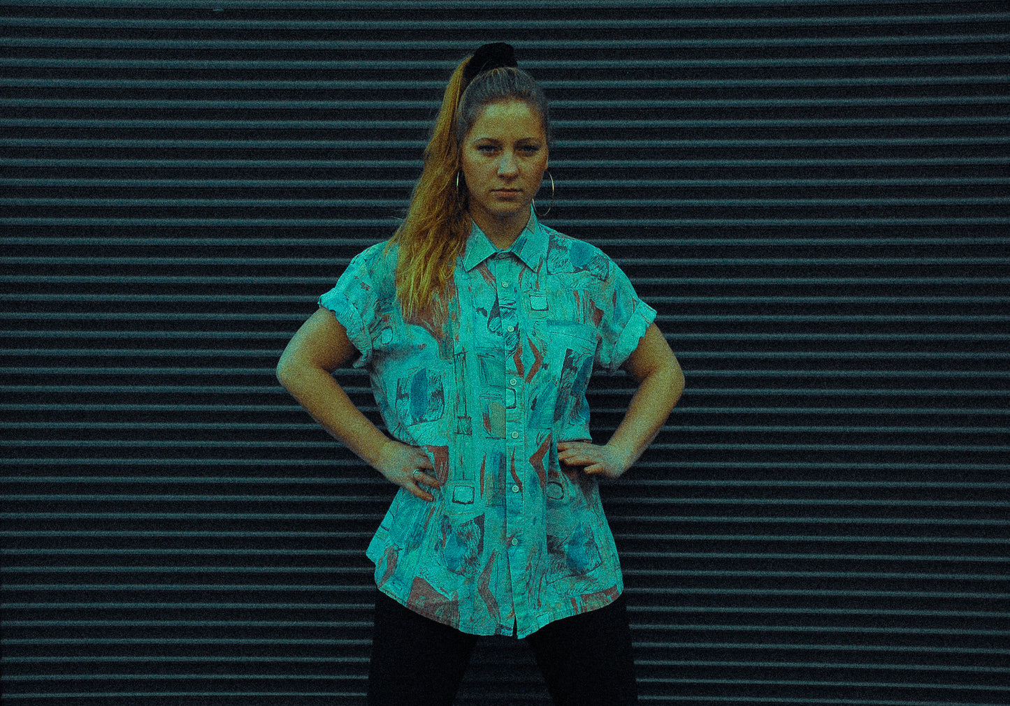 jonge-vrouw-voor-garagepoort-poserend-in-blauw-abstract-tweedehands-hemd-met-grote-gouden-ringvormige-oorbellen-en-zwarte-velvet-scrunchie