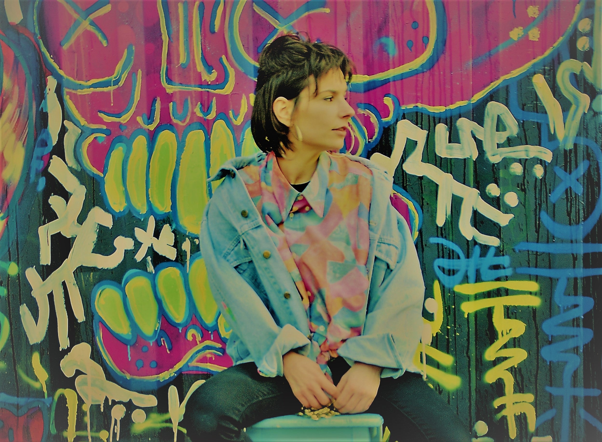 jonge vrouw draagt jaren 90 vintage kleding waaronder een abstract vintage hemd een baggy vintage jeansjas en ronde gouden oorbellen zittend voor graffitimuur